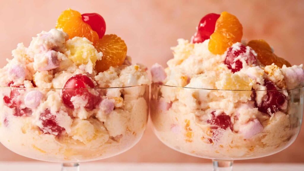Two glass dessert bowls filled with ambrosia salad made of whipped cream, marshmallows, cherries, mandarin oranges, and pineapple chunks.