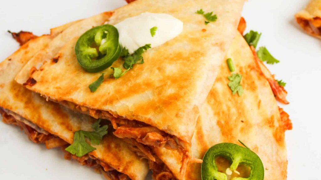 A stack of quesadillas with melted cheese and shredded meat, garnished with sliced jalapeños, chopped cilantro, and a dollop of sour cream on a white plate.