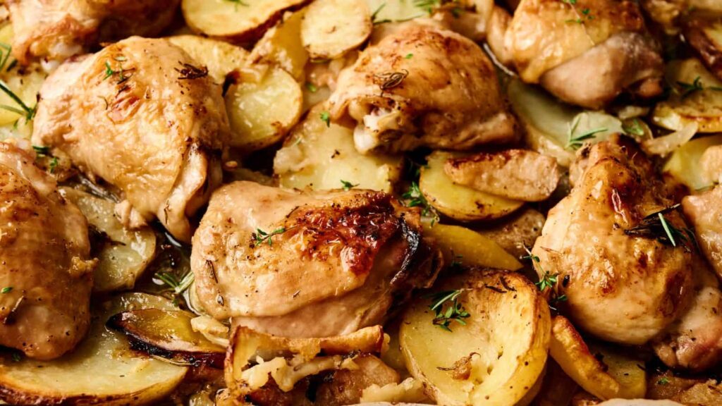 Roasted chicken thighs with slices of potatoes and herbs on a baking sheet.