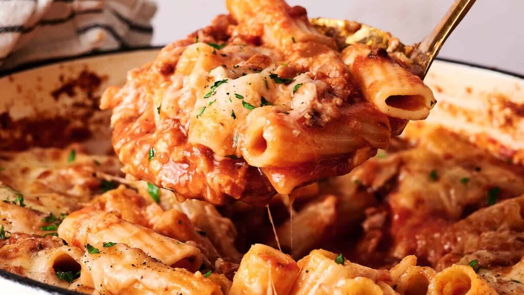 A spoon scoops baked ziti pasta covered in melted cheese and tomato sauce from a casserole dish.
