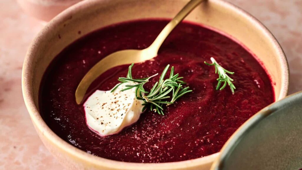 A bowl of beet soup is garnished with a dollop of cream, black pepper, and fresh rosemary, with a gold spoon resting inside the bowl.