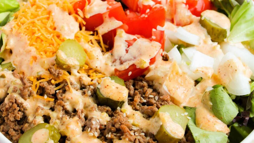 A close-up of a salad with seasoned ground beef, shredded cheese, sliced pickles, tomatoes, onions, and a creamy dressing on a bed of lettuce.
