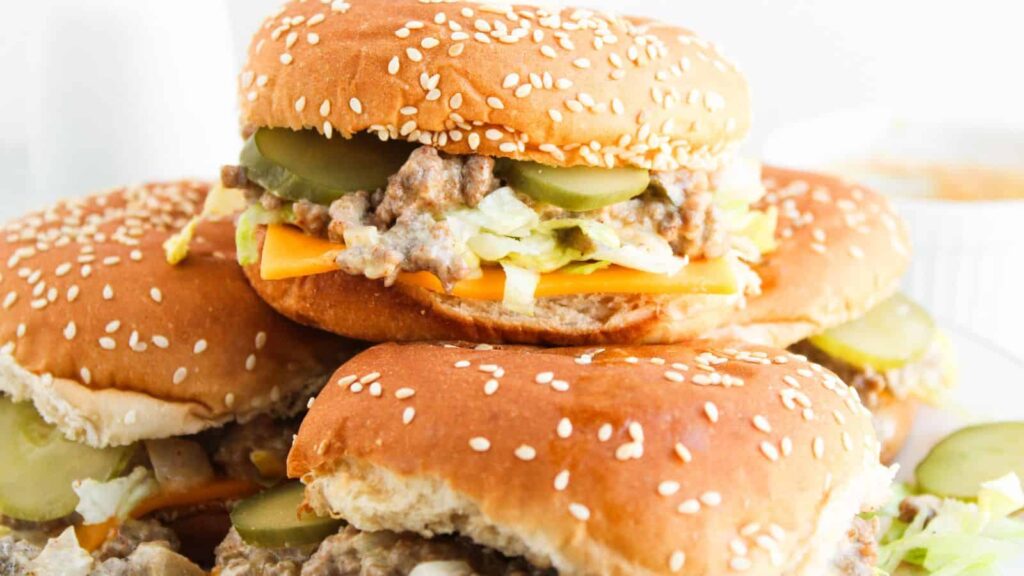 Three cheeseburgers with sesame seed buns, cheddar cheese, lettuce, pickles, and a beef patty stacked on a plate.