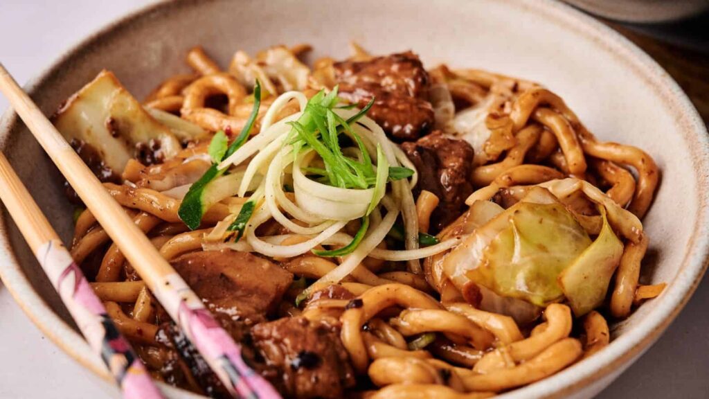 A bowl of stir-fried noodles with chunks of beef, cabbage, and green onions, served with chopsticks.