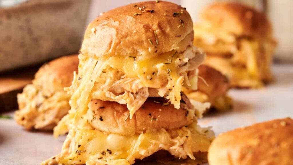 Two stacked pulled chicken and melted cheese sliders on soft buns, sprinkled with herbs, with more sliders blurred in the background.