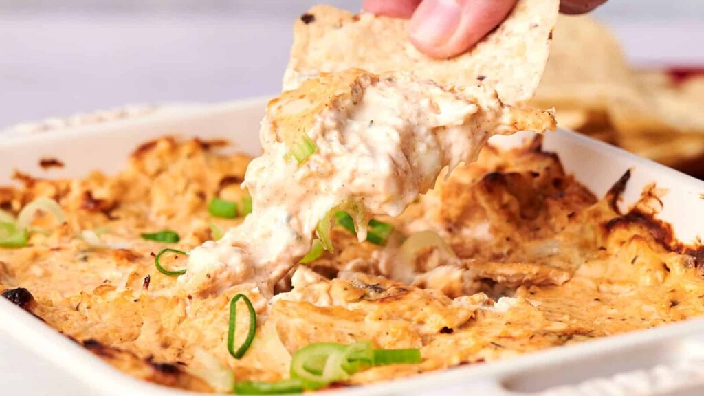 A hand scoops creamy, baked dip with a tortilla chip from a dish, topped with sliced green onions.