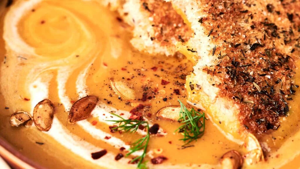 Close-up of a bowl of creamy soup garnished with pumpkin seeds, herbs, and spices. A piece of toasted bread is placed on the soup's surface.