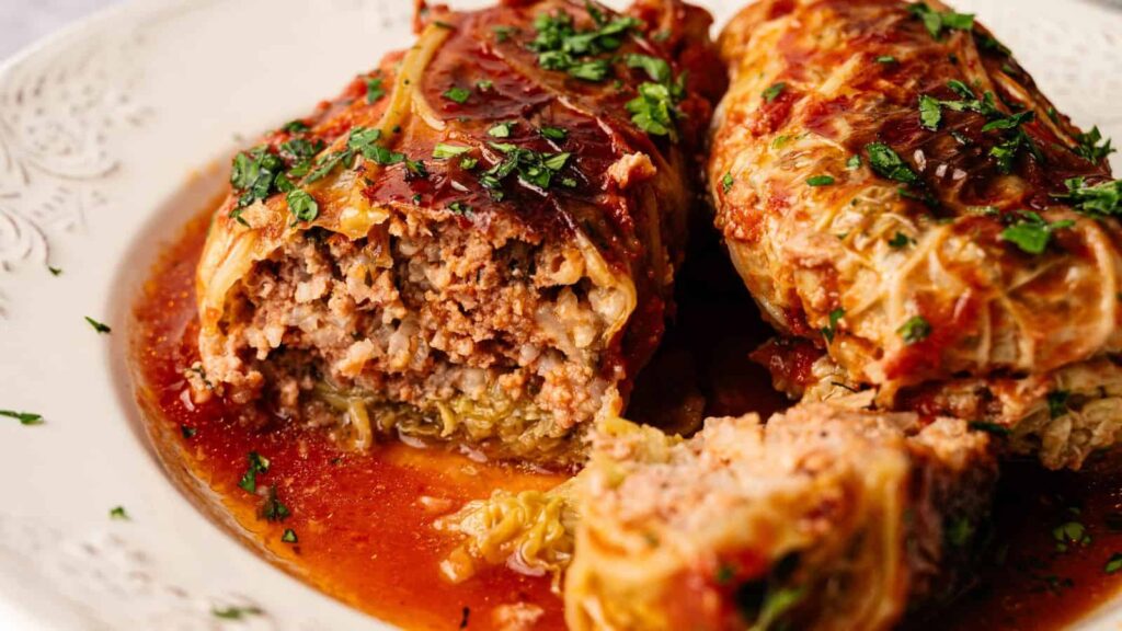 Two cabbage rolls filled with ground meat and rice, topped with tomato sauce and chopped herbs, served on a white plate.