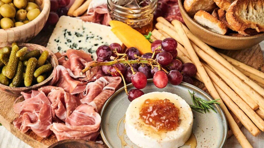 A charcuterie board with grapes, prosciutto, breadsticks, cornichons, blue cheese, a cheese round topped with marmalade, and a bowl of sliced bread.