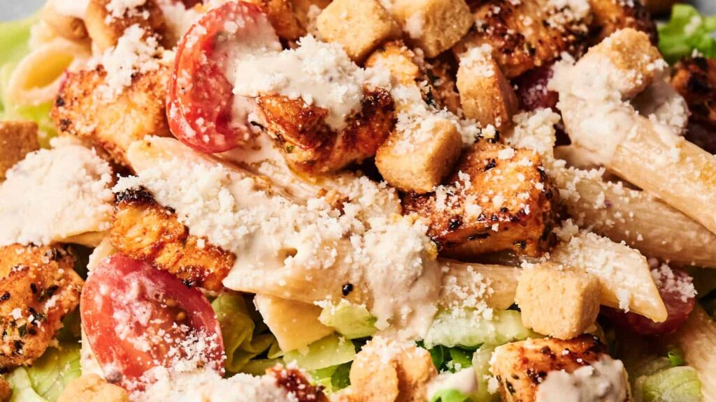 Close-up of a Caesar salad with grilled chicken, penne pasta, cherry tomatoes, croutons, lettuce, and a sprinkle of Parmesan cheese.