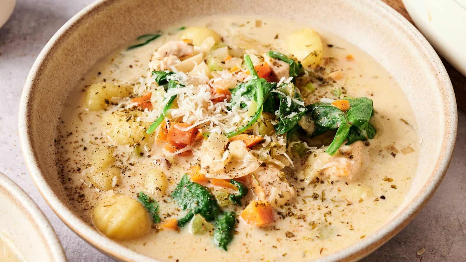A bowl of creamy soup with gnocchi, spinach, carrots, chicken pieces, and shredded cheese, garnished with herbs and black pepper.