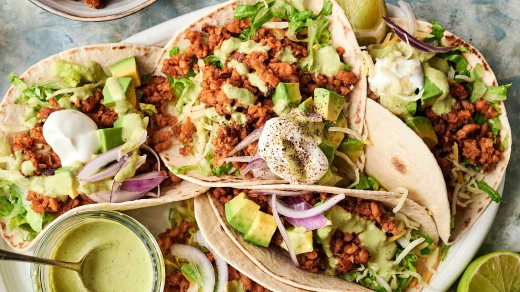 Three soft shell tacos filled with seasoned ground meat, shredded cheese, lettuce, avocado, red onions, and topped with green sauce and sour cream, served with lime wedges.