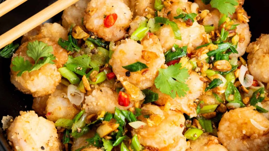 Close-up of a dish featuring fried shrimp garnished with chopped green onions, cilantro, garlic, and sliced red chili peppers, with chopsticks on the side.