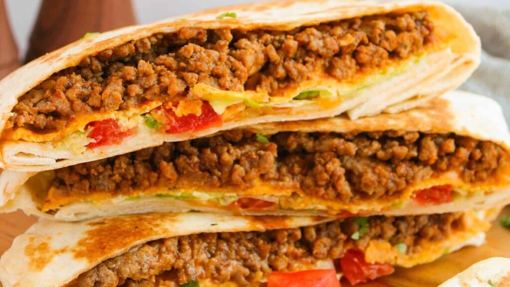 Close-up of folded tortillas filled with seasoned ground meat, melted cheese, and diced tomatoes, stacked on a wooden surface.