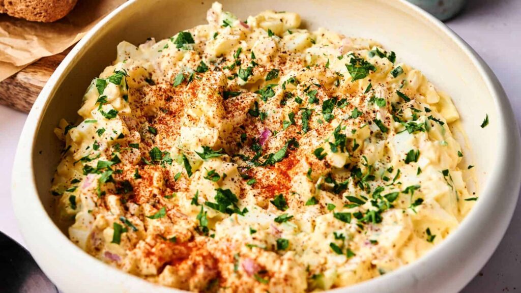 A bowl of creamy egg salad garnished with chopped parsley and paprika.