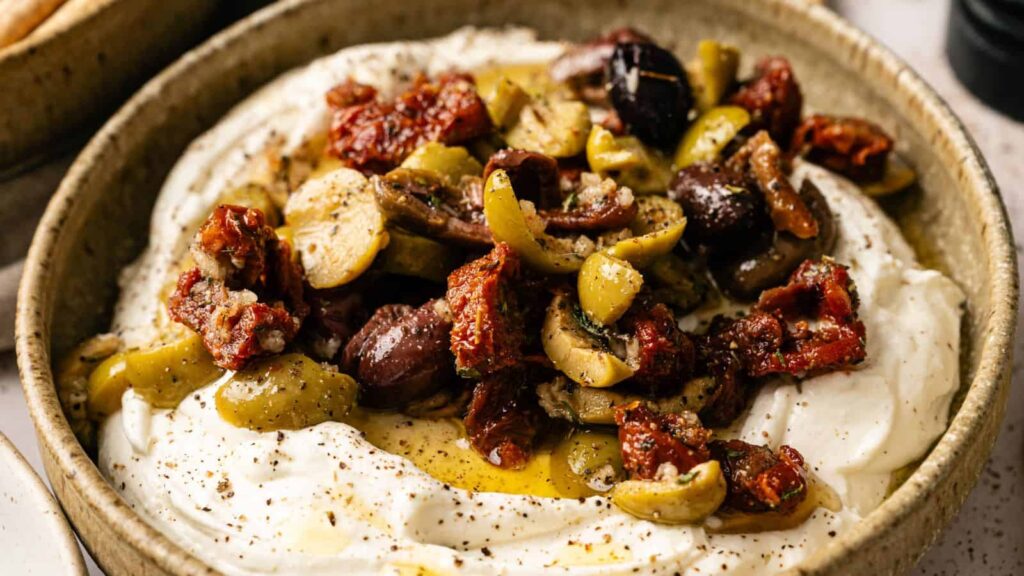 A bowl of whipped goat cheese topped with mixed olives, sun-dried tomatoes, herbs, and olive oil.
