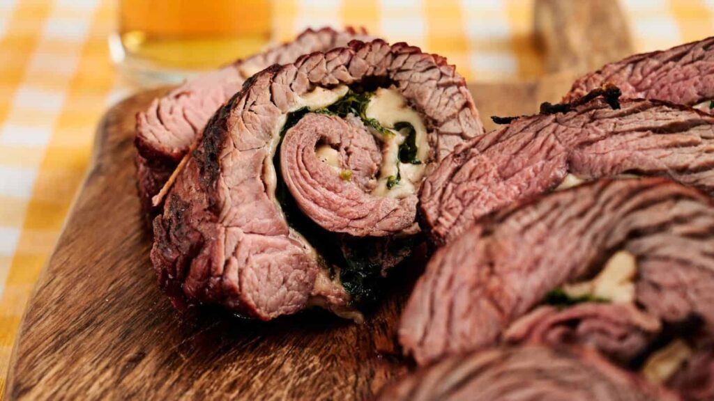 Close-up of sliced rolled beef stuffed with cheese and greens on a wooden cutting board, with a drink in the background.