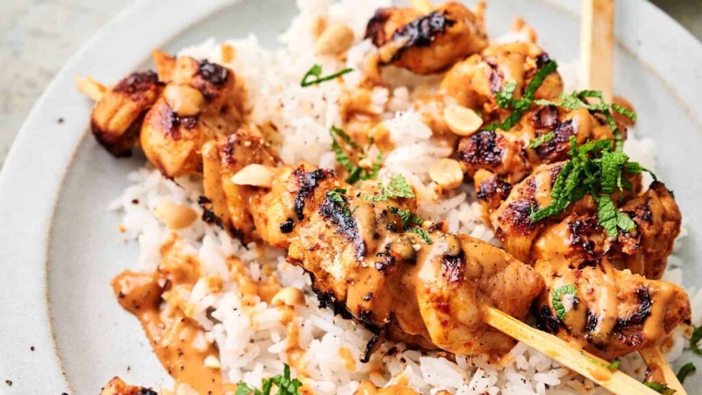Chicken skewers drizzled with peanut sauce served over white rice, garnished with chopped peanuts and fresh herbs.