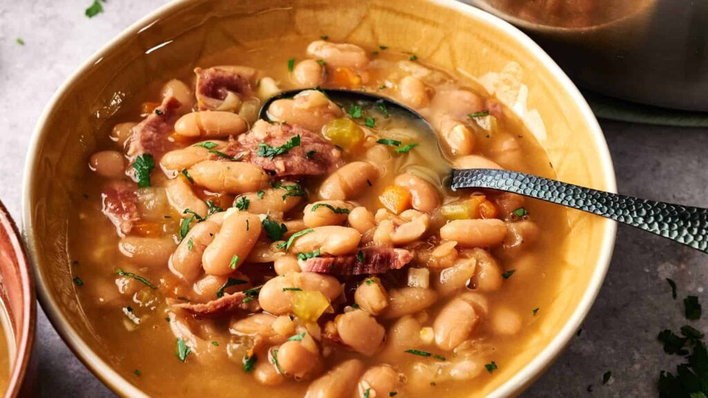 A bowl of white bean soup with pieces of ham, garnished with chopped parsley, and a spoon resting in the bowl.