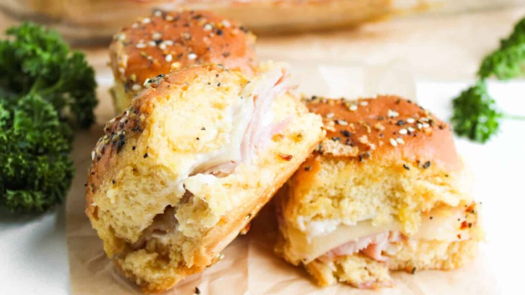 Two ham and cheese sliders on a cutting board, topped with seasoning. Parsley garnish in the background.