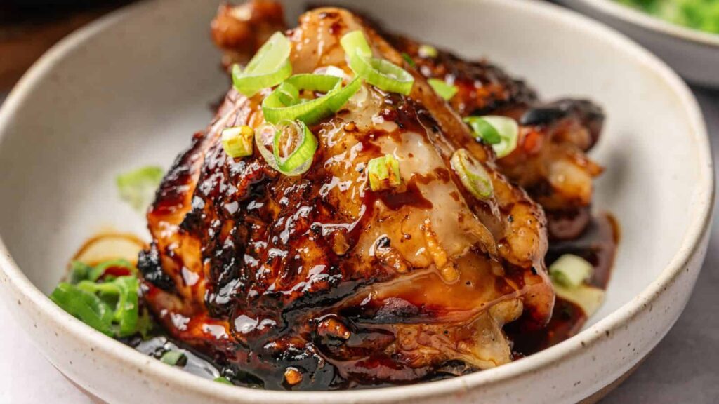 A bowl with glazed, grilled chicken topped with sliced green onions and sauce, served in a white dish.