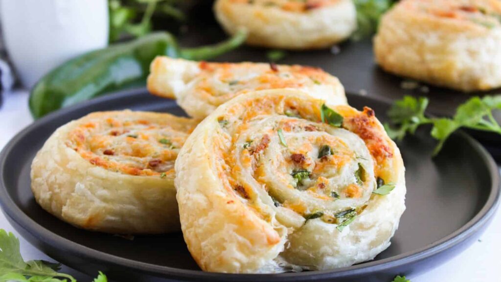 Close-up of two puff pastry spirals filled with cheese and herbs on a black plate, garnished with fresh parsley.