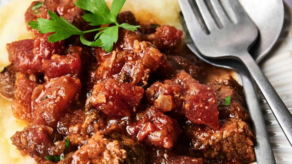 Close-up of a plate with beef stew in tomato sauce, garnished with parsley, served on a bed of mashed potatoes with a fork and spoon beside it.