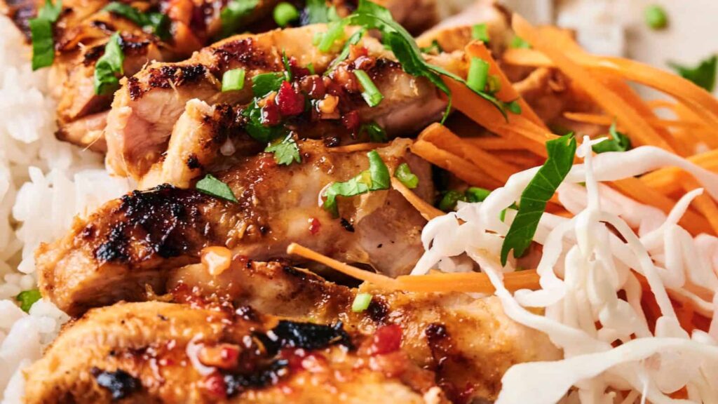 Close-up of grilled chicken slices topped with herbs and sauce, served on white rice, alongside shredded carrots and cabbage.