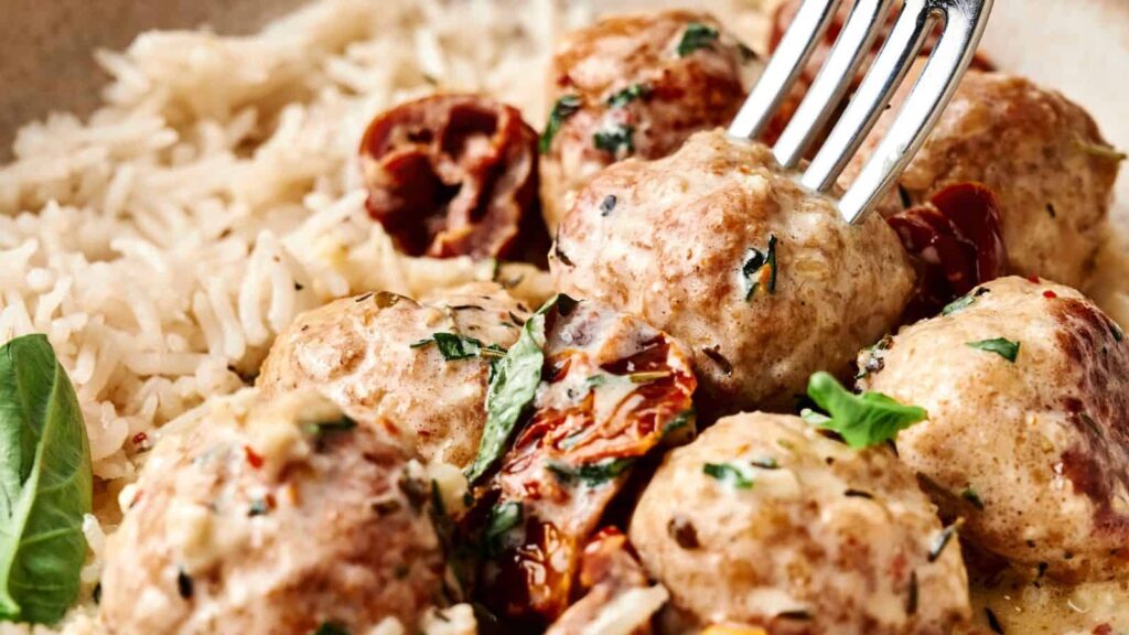 A close-up of meatballs in a creamy sauce, garnished with herbs and served with white rice and sun-dried tomatoes. A fork is inserted into one meatball.