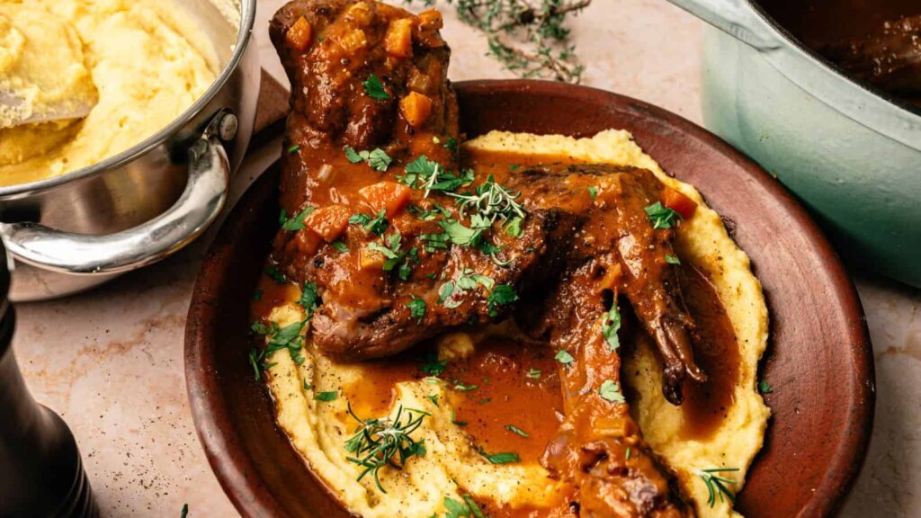 A plate of braised lamb shanks in a tomato-based sauce served over creamy polenta, garnished with fresh herbs.