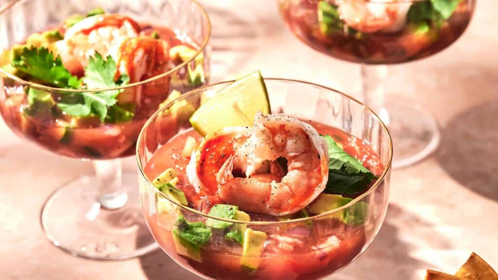 Three glasses of shrimp cocktail with avocado, lime wedges, and cilantro, served in a tomato-based sauce.