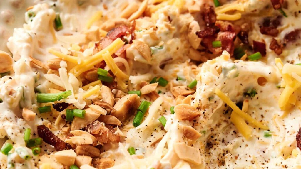 Close-up of a creamy dip topped with shredded cheese, chopped chives, sliced almonds, bacon pieces, and ground black pepper.
