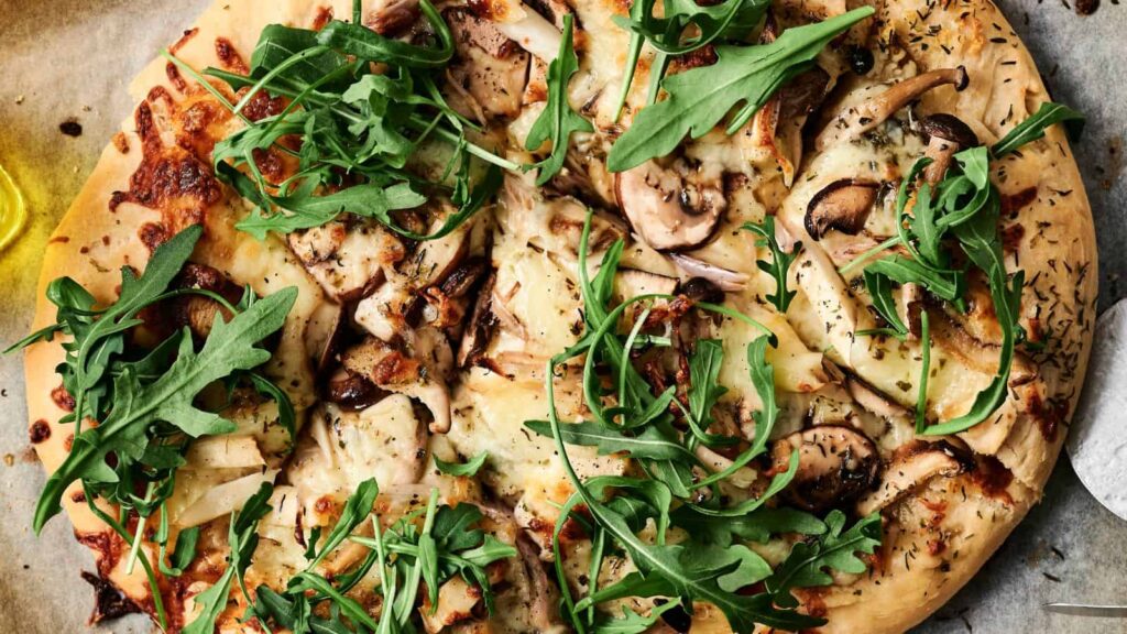 Close-up of a pizza topped with mushrooms, cheese, and fresh arugula leaves on a wooden surface.