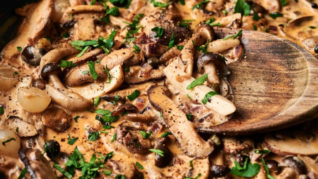 Creamy mushroom stew with sliced mushrooms, herbs, and black pepper, stirred with a wooden spoon.