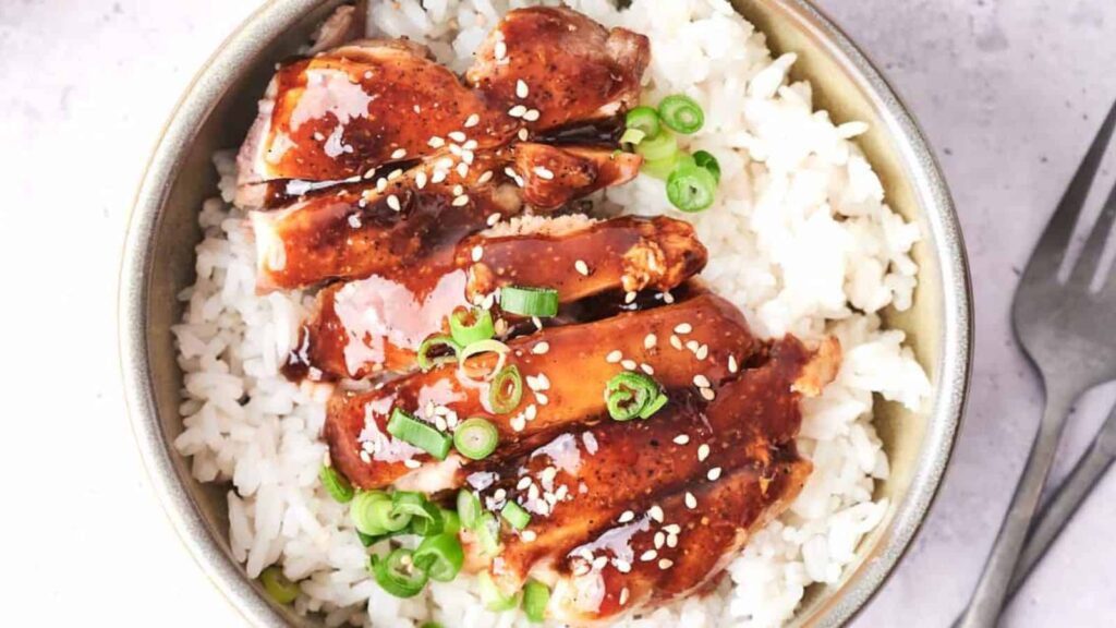 Bowl of rice topped with glazed chicken and garnished with green onions and sesame seeds.