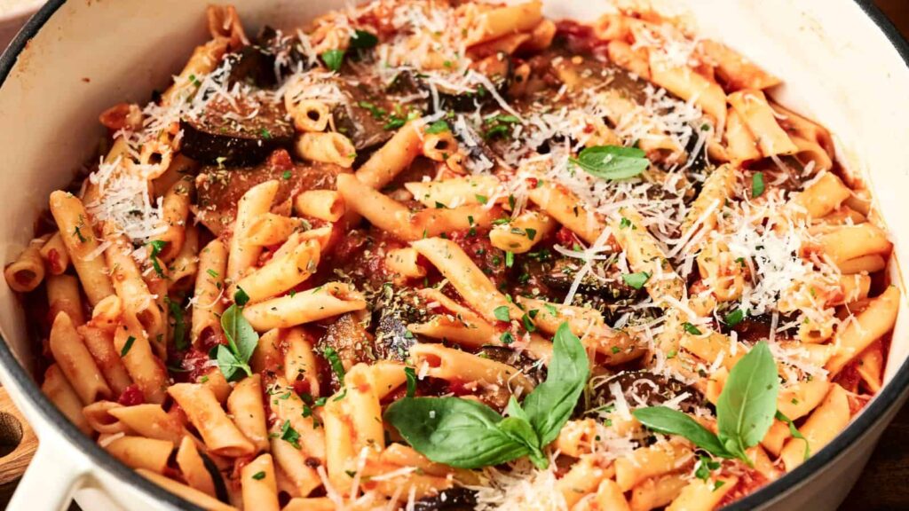 A pot of penne pasta mixed with tomato sauce, topped with grated cheese, herbs, and basil leaves.