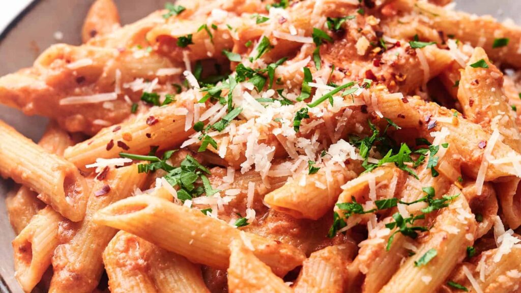 Close-up of penne pasta topped with tomato cream sauce, grated Parmesan cheese, chopped parsley, and red pepper flakes.