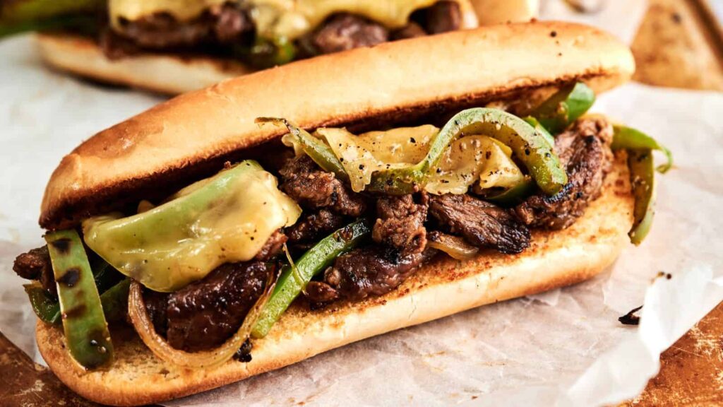 A close-up of a Philly cheesesteak sandwich filled with sliced beef, melted cheese, sautéed green bell peppers, and onions in a toasted hoagie roll.