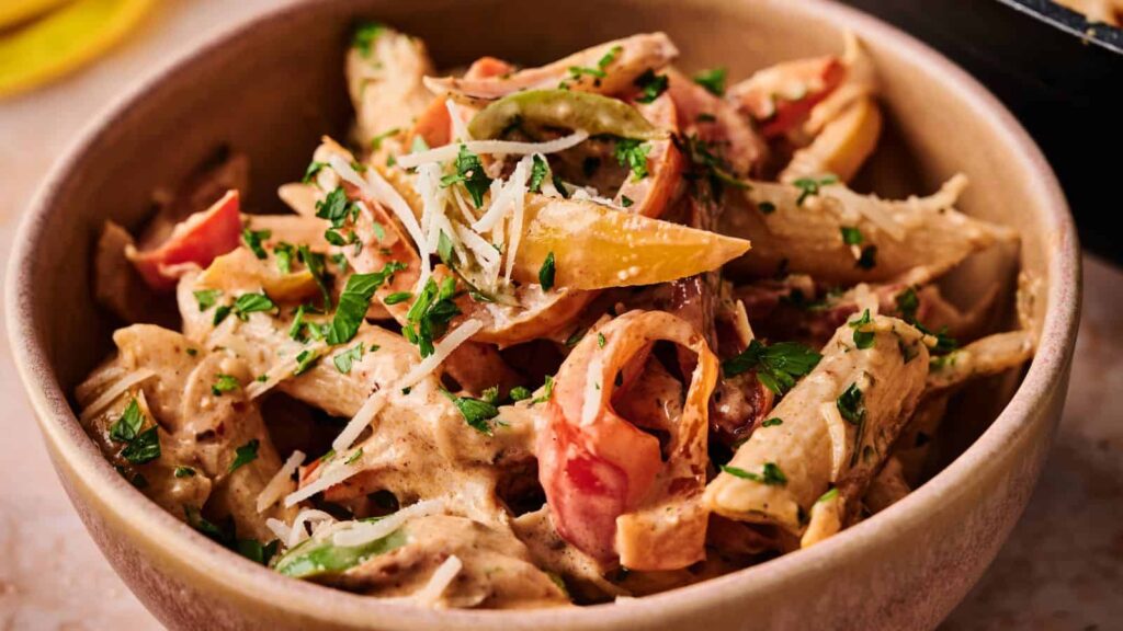 A bowl of penne pasta with creamy sauce, sliced bell peppers, and fresh parsley, topped with grated cheese.