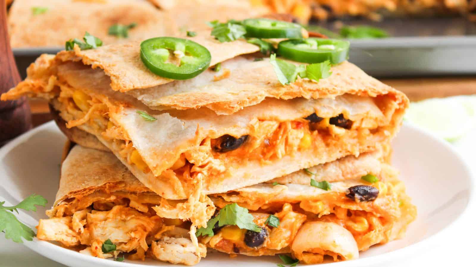 Close-up of stacked quesadilla slices filled with cheese, beans, and chicken, topped with sliced jalapeños and cilantro, on a white plate.
