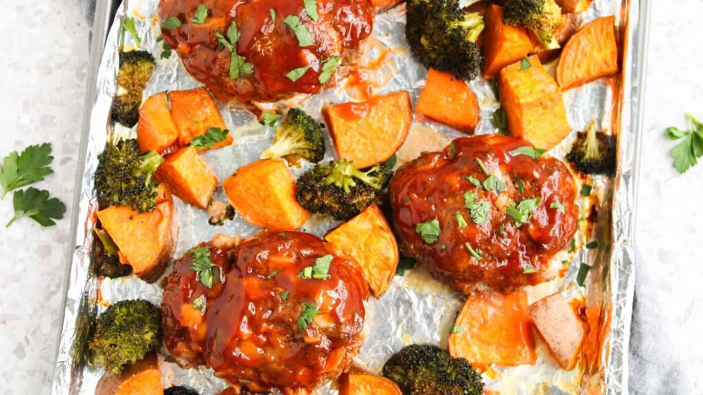 Three glazed meatloaves, roasted sweet potato chunks, and broccoli florets are arranged on a foil-lined baking sheet, garnished with chopped herbs.