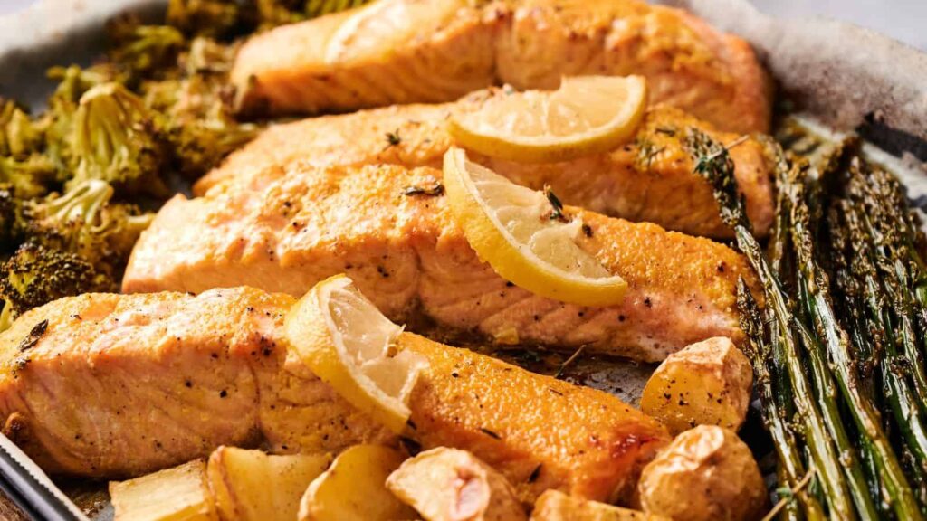 Baked salmon fillets topped with lemon slices, served with roasted broccoli, asparagus, potatoes, and garlic on a baking tray.