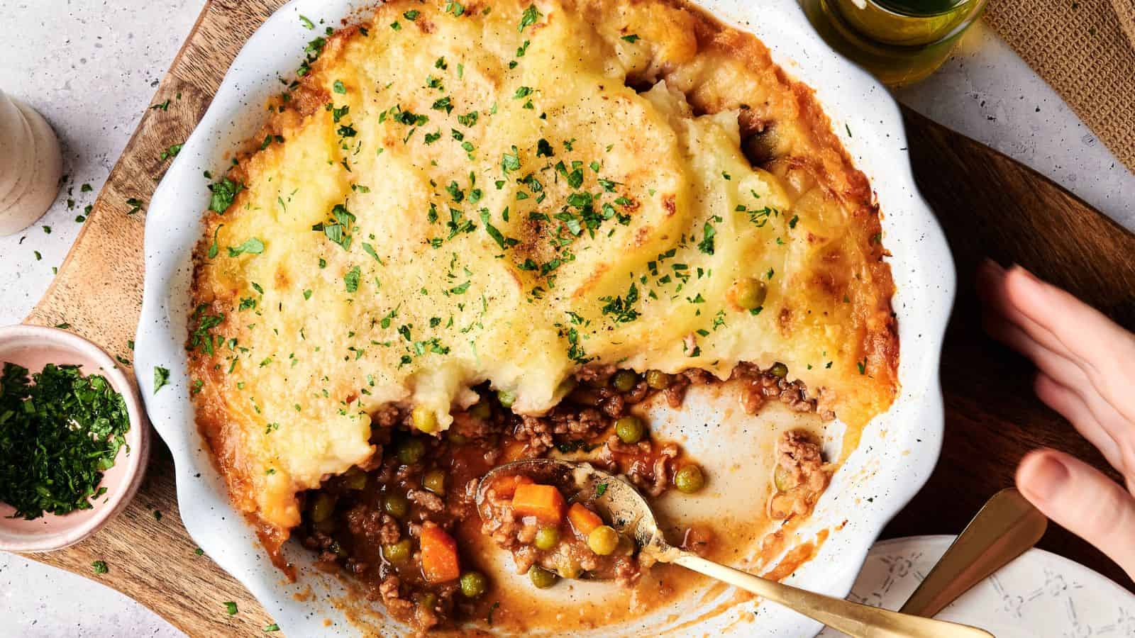 A baked shepherd's pie with a golden mashed potato topping, partially served to reveal ground meat, peas, carrots, and gravy in a white dish.