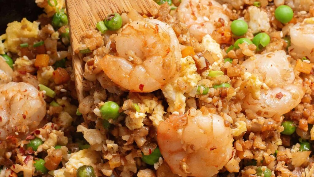 Close-up of shrimp fried rice with peas, scrambled eggs, diced vegetables, and shrimp, being stirred with a wooden spatula.