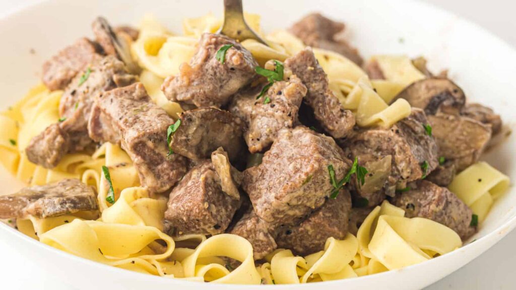 A close-up of beef stroganoff with wide egg noodles, featuring tender beef chunks in a creamy sauce garnished with herbs.