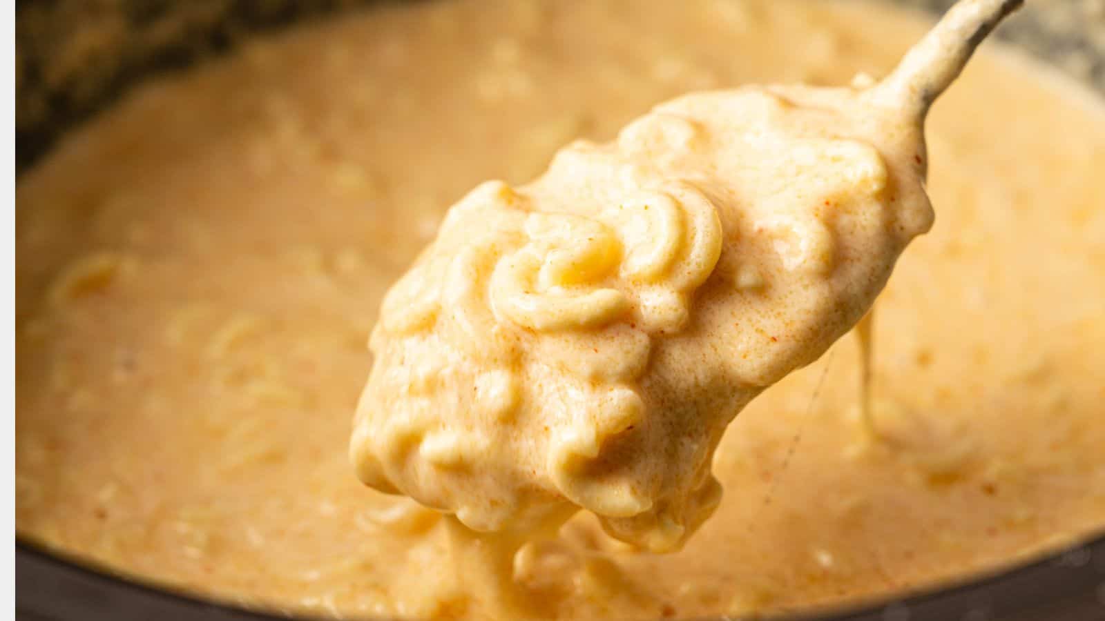 A close-up of a spoon lifting creamy, melted cheese sauce with macaroni from a pot.