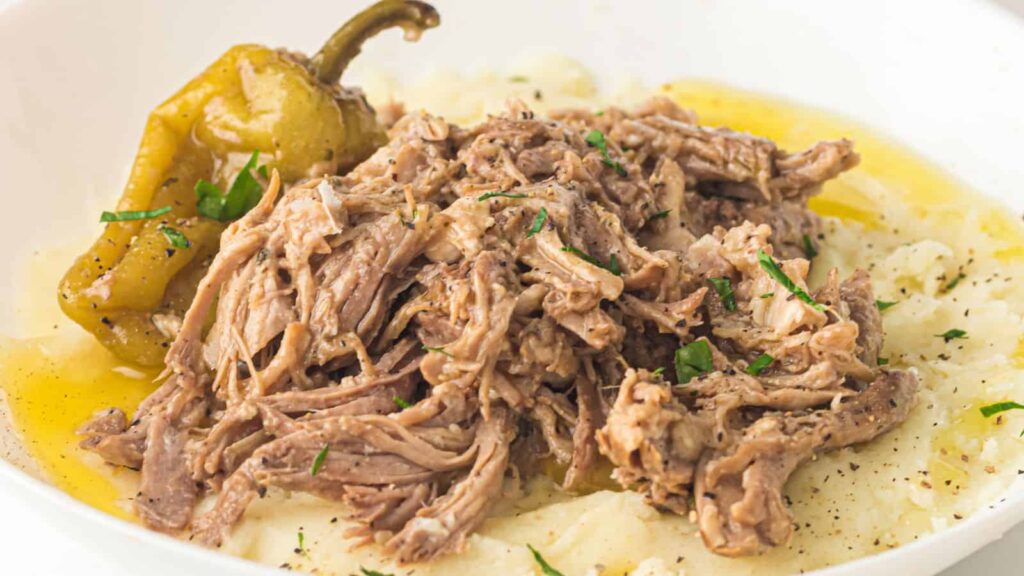 Shredded beef with herbs served over mashed potatoes, topped with a whole pepper and drizzled with broth in a white bowl.