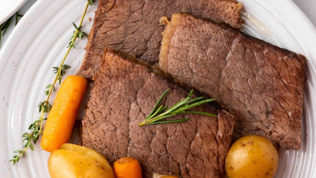 Slices of roast beef with rosemary, carrots, and potatoes on a white plate.