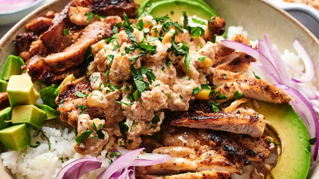 A bowl of rice topped with grilled chicken, creamy sauce, chopped cilantro, avocado slices, and red onion strips.