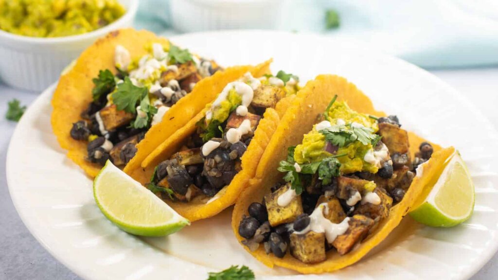 Three tacos filled with black beans, diced vegetables, sauce, and cilantro are arranged on a white plate with lime wedges on the side.